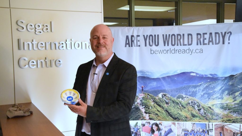 Sean Kennedy is pictured revealing the 2020 NC Diya during the filming of the video introduction. He is standing in front of a Be World Ready banner at the Segal International Centre. 