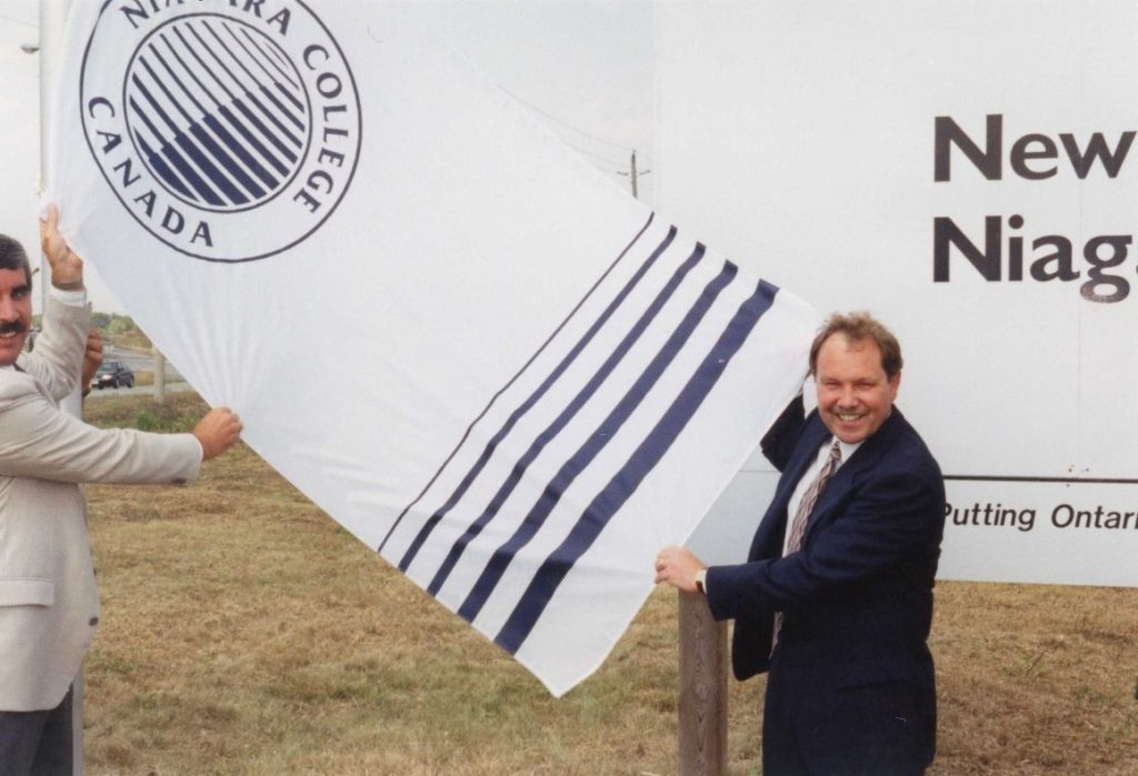 Dan Patterson holding a Niagara College flag in front of a large sign