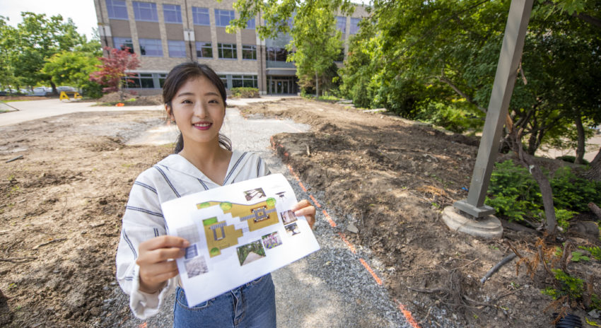 Student Alice Lee is holdiing up her design plans outside at the Daniel . Patterson Campus in Niagara-on-the-Lake where work is underway for the memorial garden.