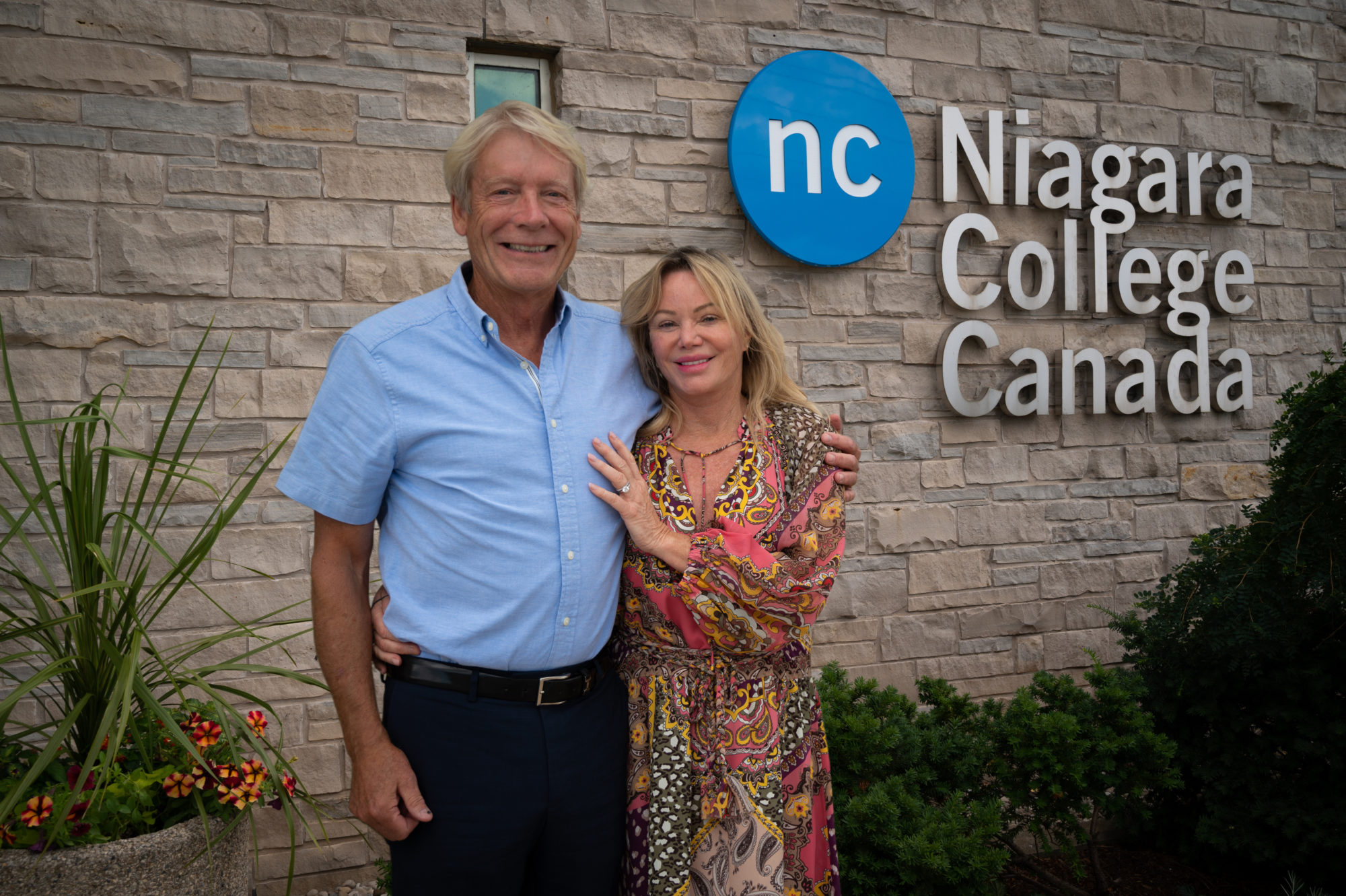 A smartly dressed couple stands in front of the a building that features the Niagara College logo.