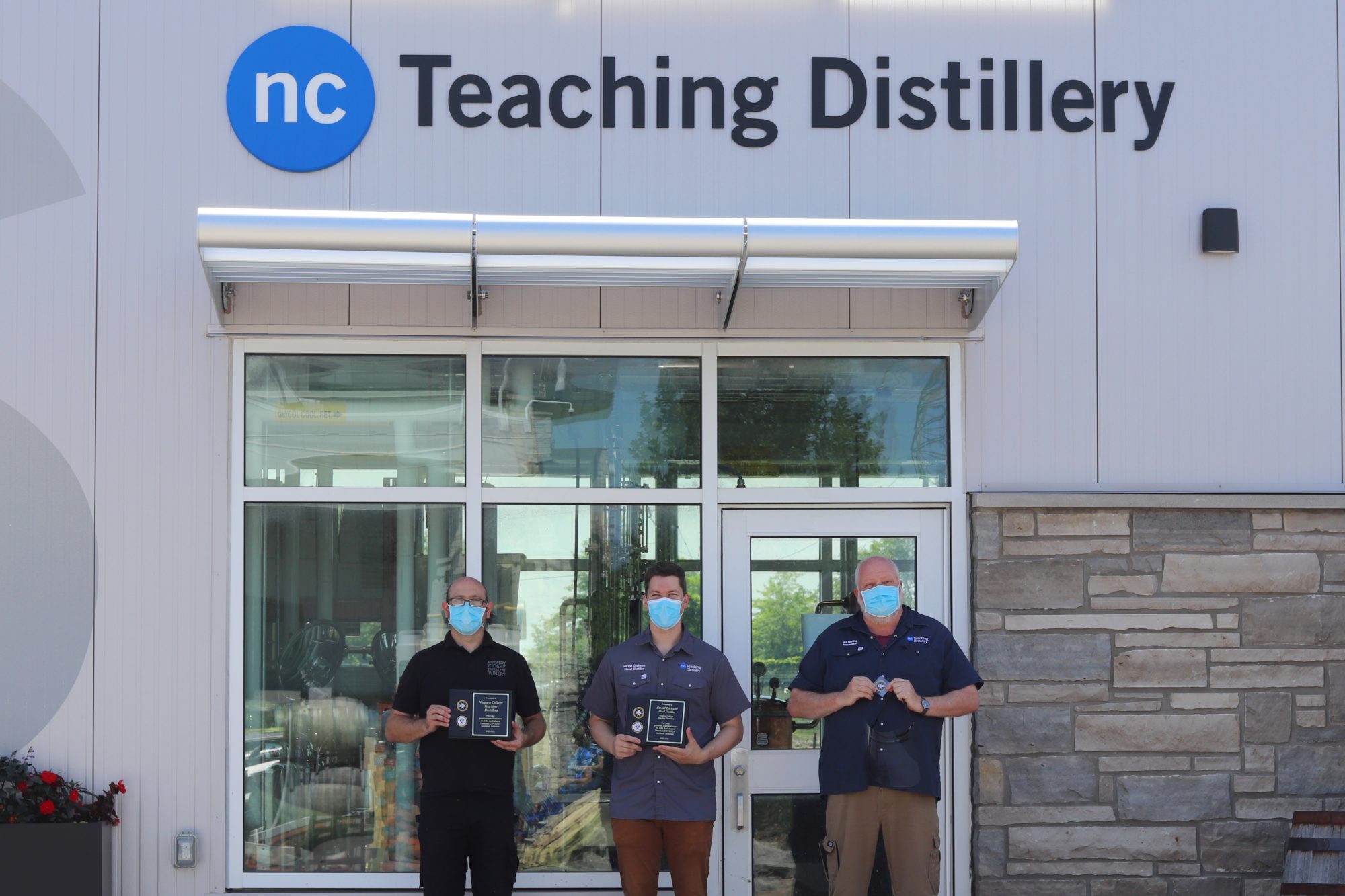 Three men stand in front of the Teaching Distillery holding placques.