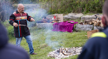 Man wearing stands before a smoking fire and addresses a crowd of people gathered at the College's Indigenous garden.