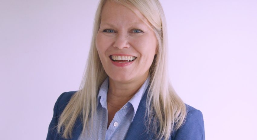 A head shot of a woman with long blonde hair wearing a blue collared shirt and suit jacket smiles at the camera