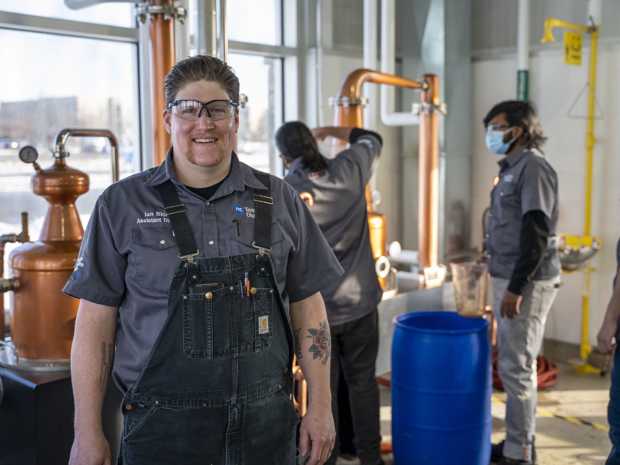 A man in overalls and safety glasses stands in the foreground while two people work on distilling equipment behind him.