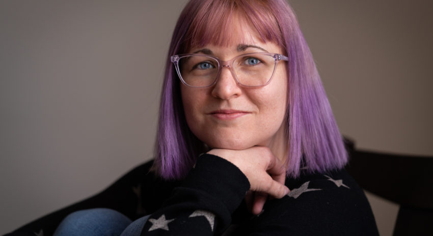 Photo of a woman in glasses with purple hair sitting with her chin resting on her hand.