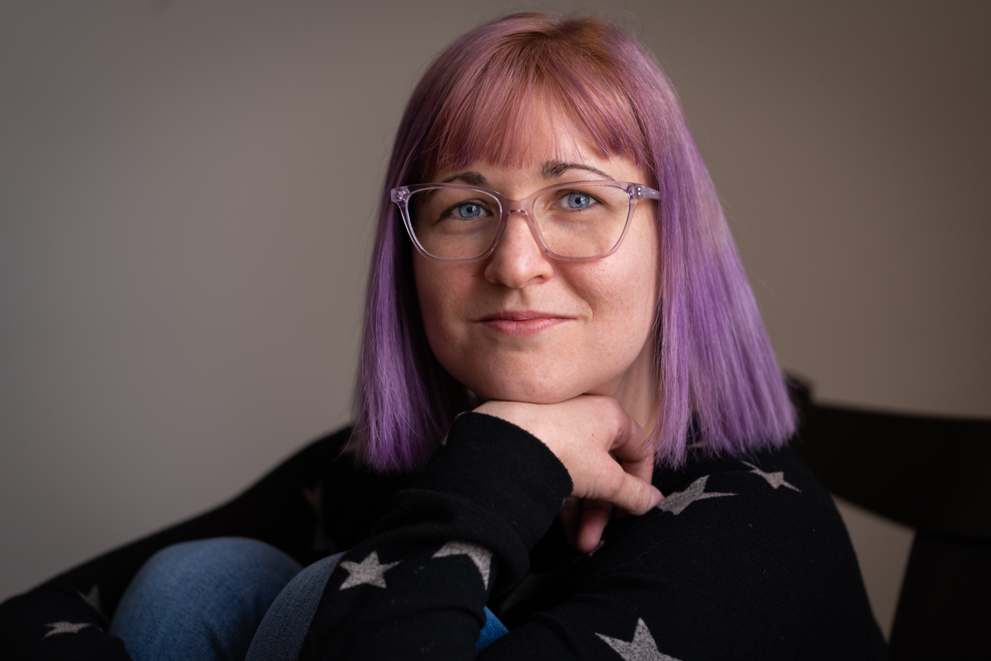 Photo of a woman in glasses with purple hair sitting with her chin resting on her hand.
