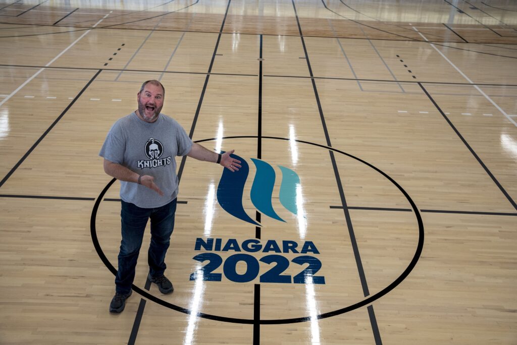a man wearing a grey t-shirt and jeans stands on a basketball court pointing to centre court