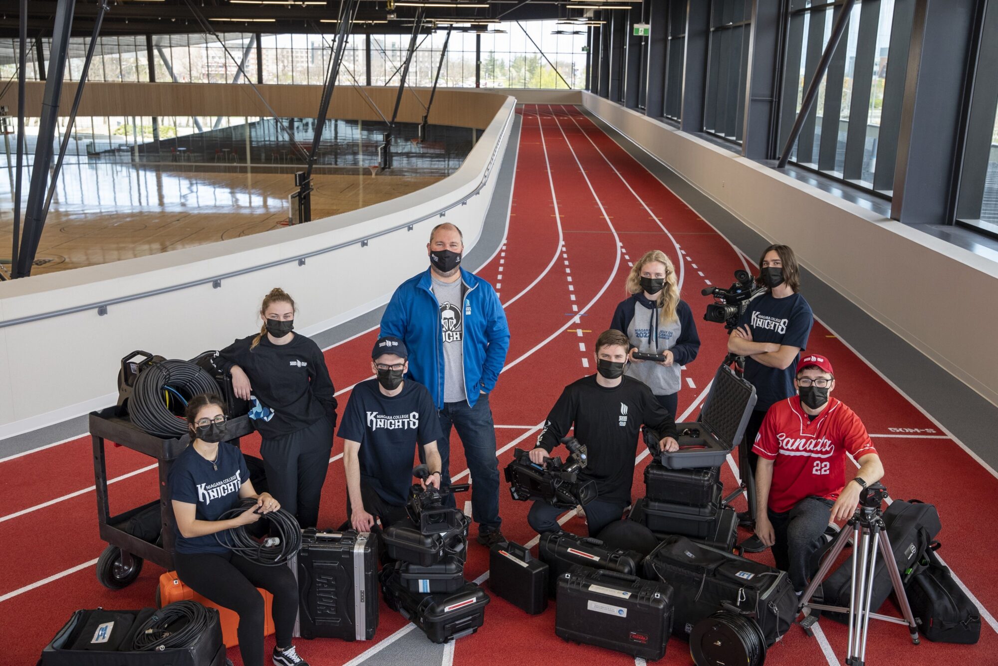 a group of 8 people stand together on an indoor sports track with production equipment